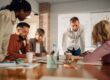Team of five collaborating around a table; one man stands and points at documents as others take notes.